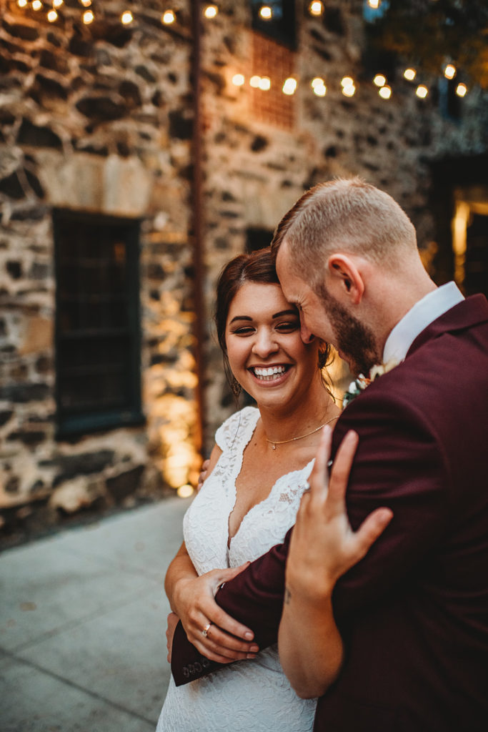 Baltimore wedding photographer captures bride and groom posing together outside of their Baltimore wedding venue with twinkle lights overhead with groom in a red suit and bride in a lace dress