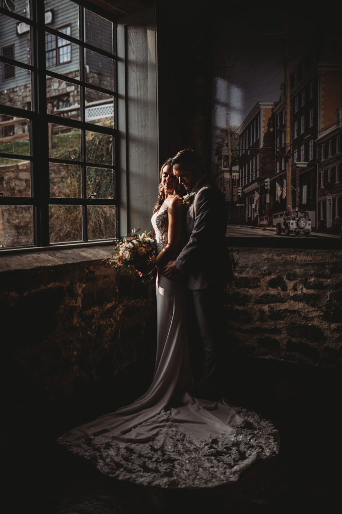 Baltimore wedding photographers captures groom embracing his bride as he stands behind her and they both look out a window together in their Baltimore wedding venue