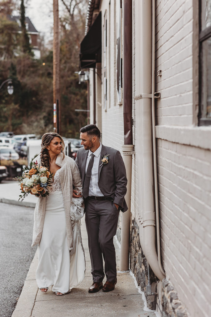 bride in a lace wedding dress and a faux fur coat holding hands with her groom who is in a grey suit as they walk down and alley in downtown Ellicott City capture by Maryland wedding photographer 