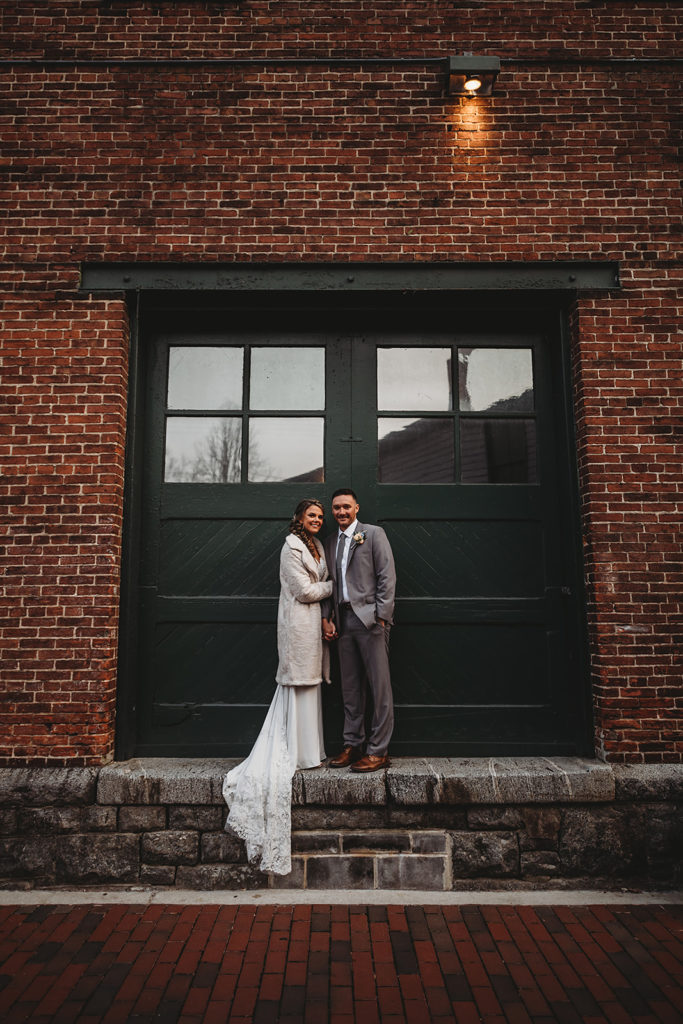Maryland wedding photographer captures winter wedding with bride and groom posing in front of a large black door on a red brick building for their timeless wedding portraits