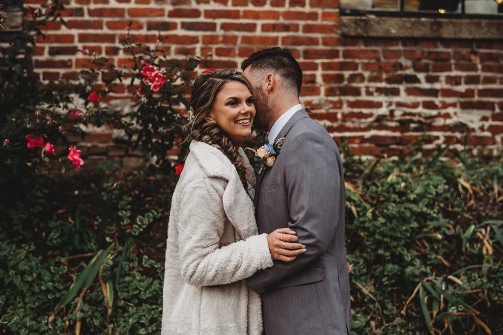 bride and groom embracing with groom whispering in his brides ear as he holds her waist with a brick building behind them with a rose garden 
