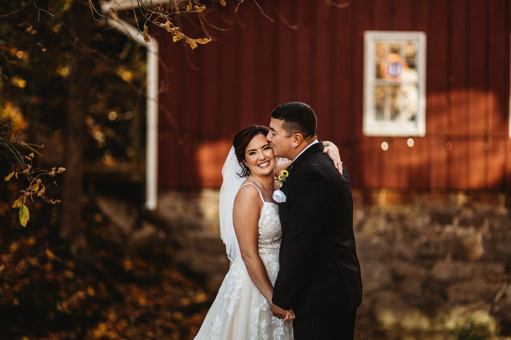 bride holding one arm around her groom's shoulder while she holds his other hand and smiles as he kisses her on the cheek captured by Maryland wedding photographer