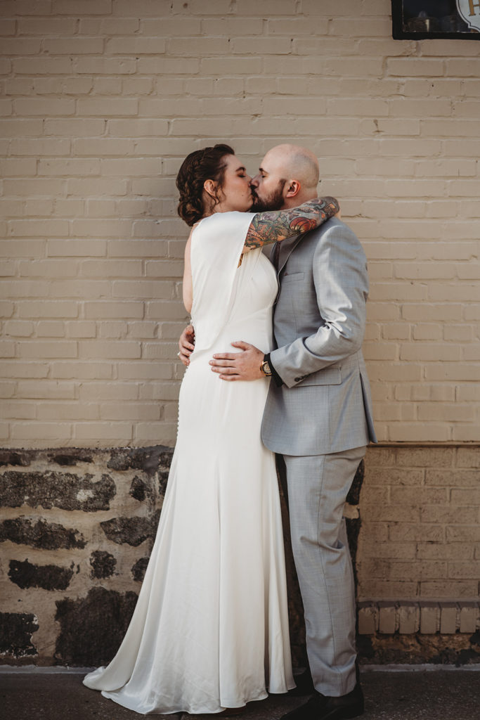 bride and groom kissing in an alley in Ellicott City with bride in a satin boho wedding dress and groom in a gray suit captured by Baltimore wedding photographers 