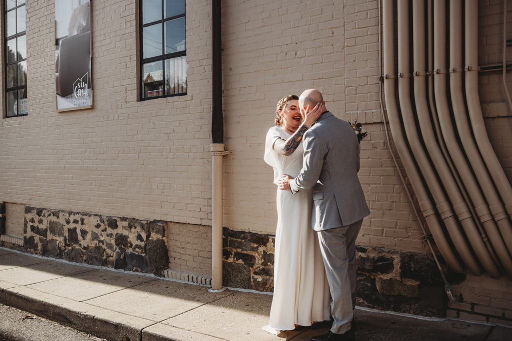 Ellicott City wedding venues with bride and groom standing outside Main Street Ballroom and embracing each other next to a white brick wall captured by Baltimore wedding photographers 