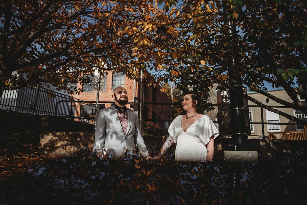 Baltimore wedding photographers captures bride and groom holding hands and standing under fall trees for their Main Street Ballroom Ellicott City wedding venues