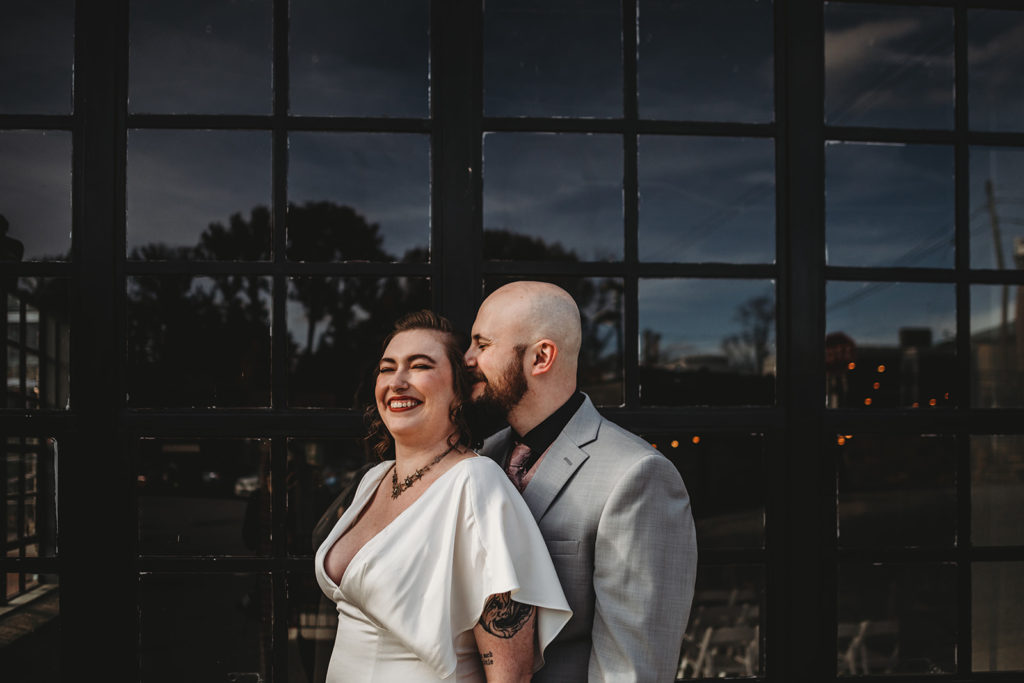 bride standing in front of her groom as he leans into her ear and whispers to her causing her to laugh as they stand in front of a large window outside of their wedding venue captured by Baltimore wedding photographer 
