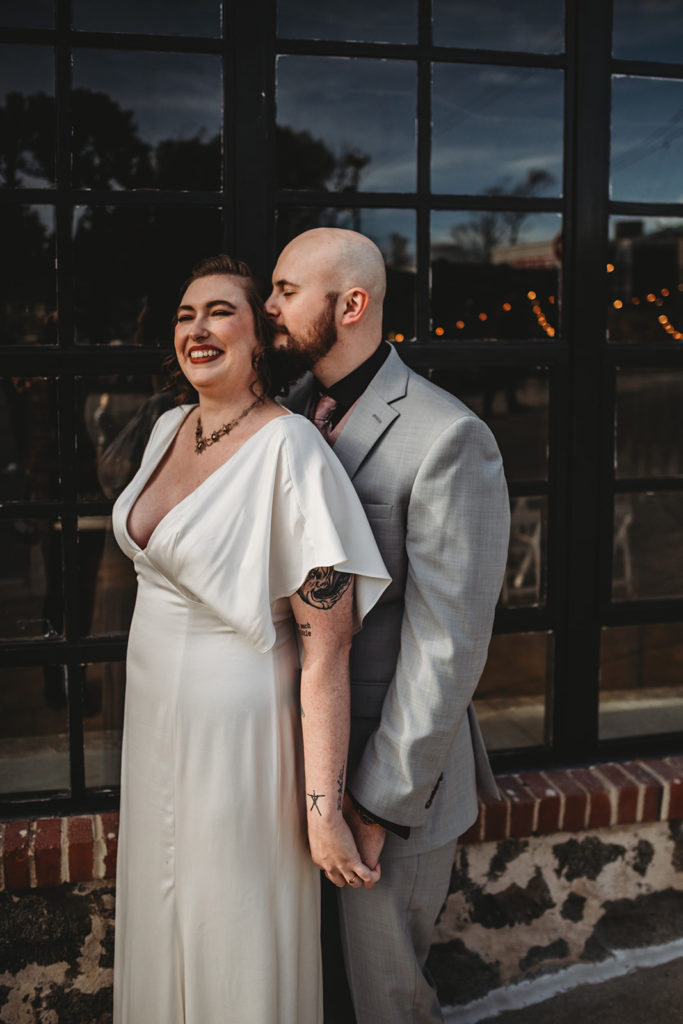 groom standing behind his bride and whispering in her ear as she leans back into him and smiles as they stand in front of a venue front captured by Baltimore wedding photographers