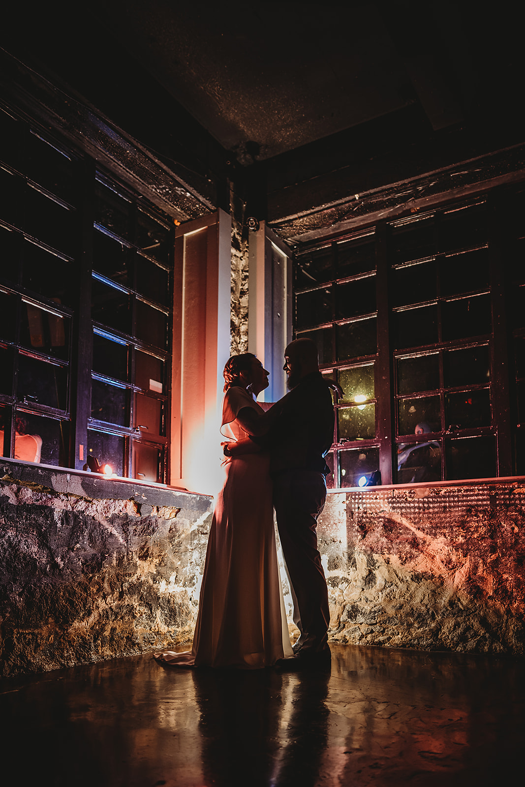 Baltimore wedding photographers captures night time wedding photography with bride adn groom embracing in a stone house with industrial windows while purple and red light shines behind them
