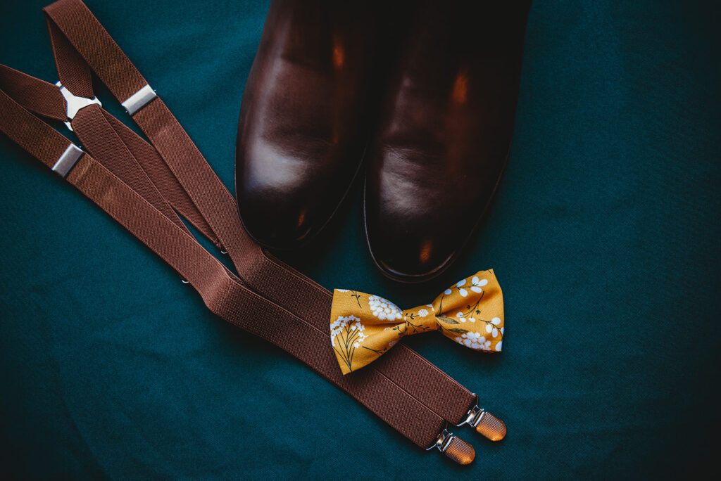 grooms brown shoes sitting on a teal cloth with his brown suspenders and yellow bowtie captured by Baltimore wedding photographers