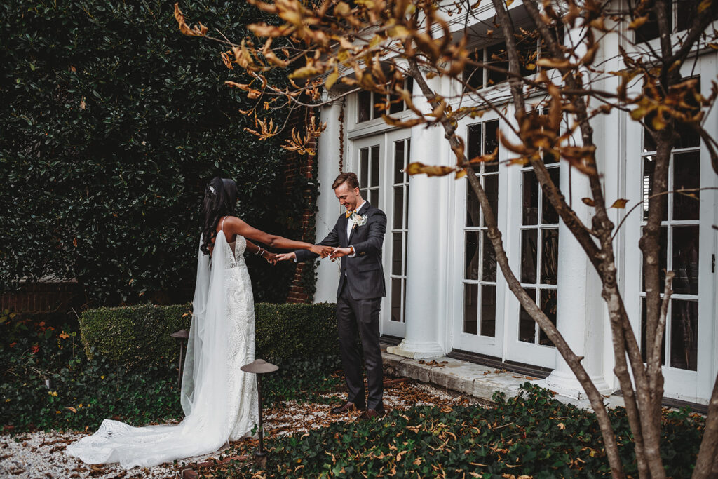 Baltimore photographers captures first luck on the wedding day between bride and groom while the groom holds the brides hands and looks at her gown in a courtyard of Belmont country club