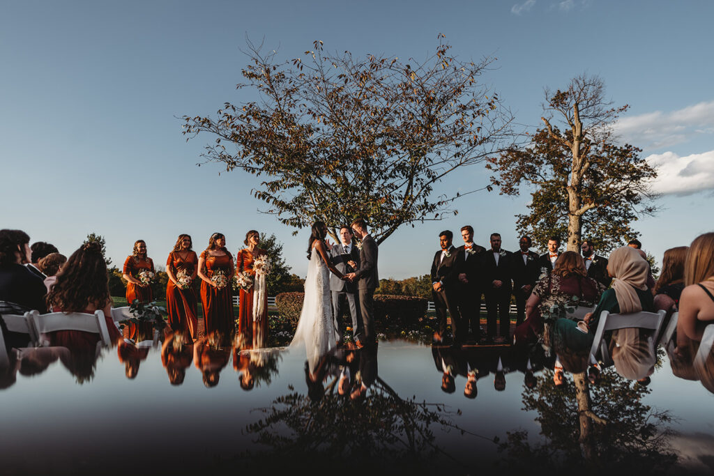 Baltimore photographers photographs outdoor wedding ceremony with bride and groom holding hands in front of a tree and the bridal party surrounding them on either side with a reflective edit