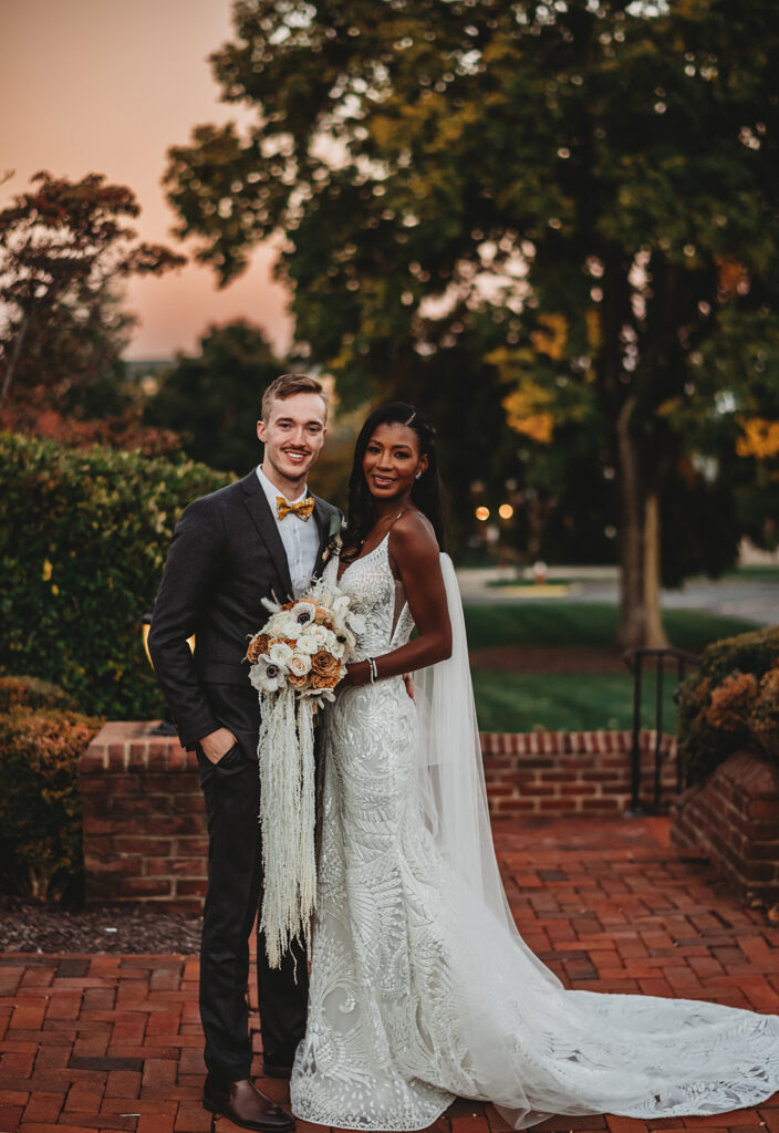 Baltimore photographers photographs sunset wedding photos with bride and groom standing on a brick porch surrounded by greenery as they pose