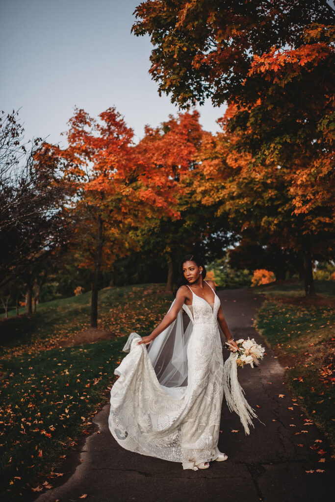 Baltimore photographers captures outdoor bridal photos with black bride in a lace wedding gown holding the train and twirling around on a path with fall leaves in the distance