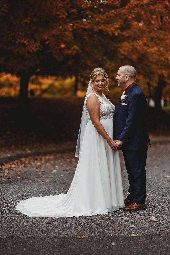 Baltimore wedding photographers photographs bride and groom holding hands while standing on a long driveway together as the fall trees stand behind them
