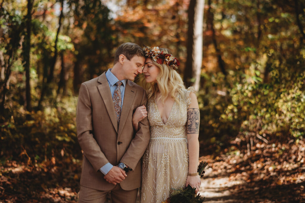 Maryland wedding photographer captures bride and groom standing side by side in the woods in the fall months while the bride holds the grooms arm and they lean in to rest their foreheads together