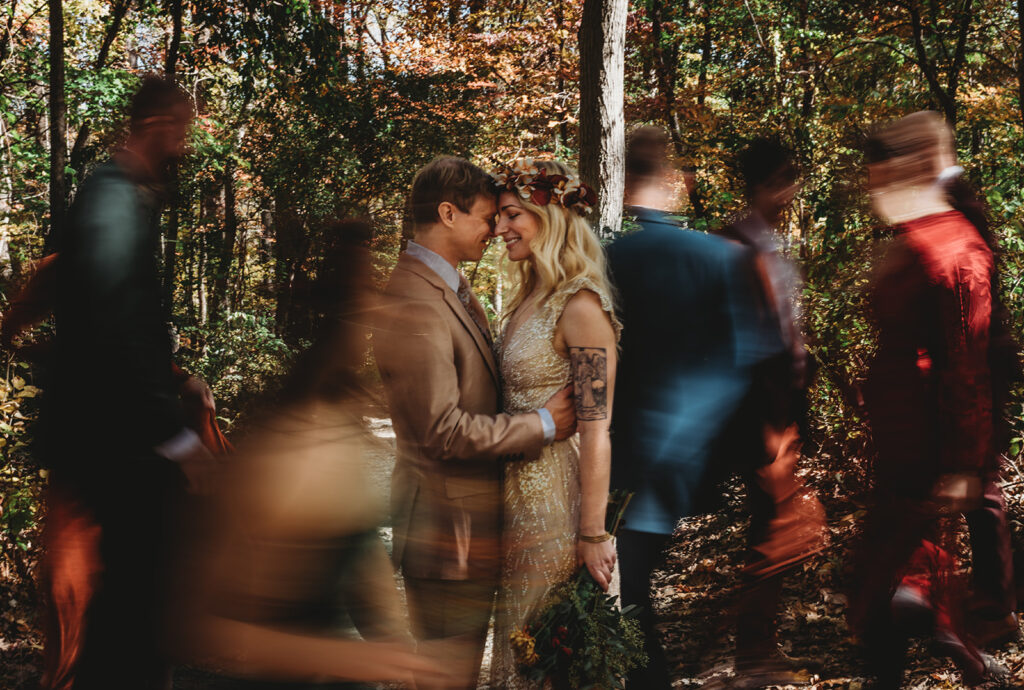 Maryland wedding photographer photographs unique wedding picture with bride and groom embracing each other in the woods with their bridal party moving around them in a blur