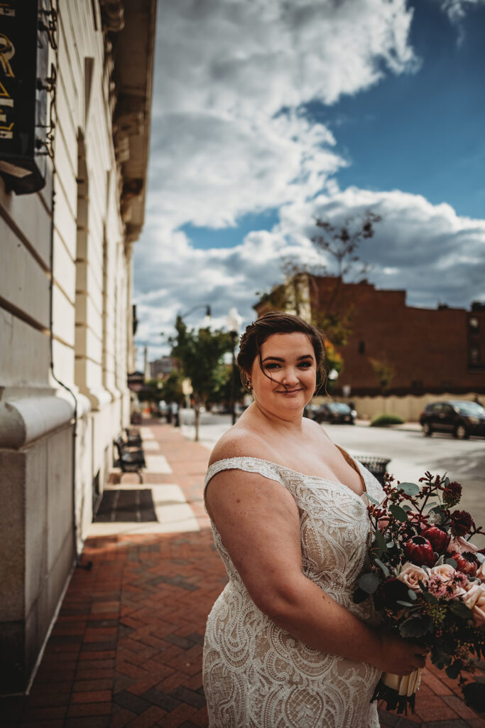 bride standing in downtown Baltimore with her burgundy florals while walking on a brick sidewalk and looking over her shoulder captured by Maryland wedding photographer