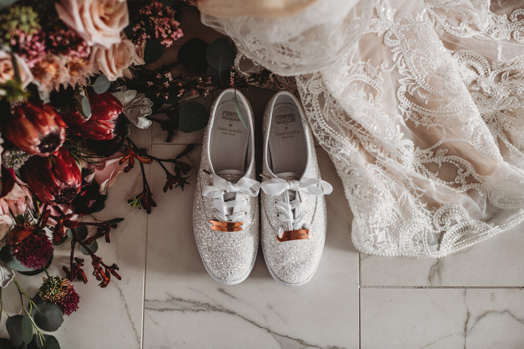 sparkly keds with wide white ribbons for bridal shoes sitting next to the brides lace wedding dress and floral bouquet