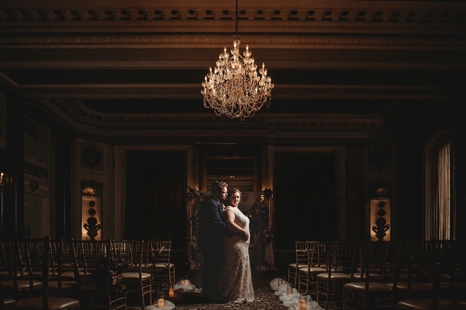 Baltimore photographers photographs bridal portraits at the Belvedere wedding venue with groom embracing bride as he stands behind her in their ceremony hall with a chandelier lit above head