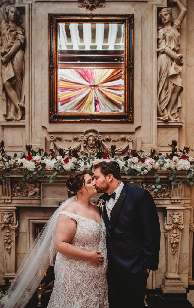Baltimore photographers photographs bridal portraits with bride and groom kissing in front of a marble fireplace decorated with roses