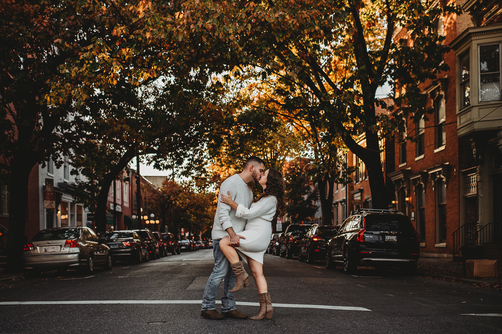 Baltimore photographers captures fall engagement pictures in Baltimore with man and woman kissing in the middle of a road in downtown with historic building lining the streets with old trees hanging over the road