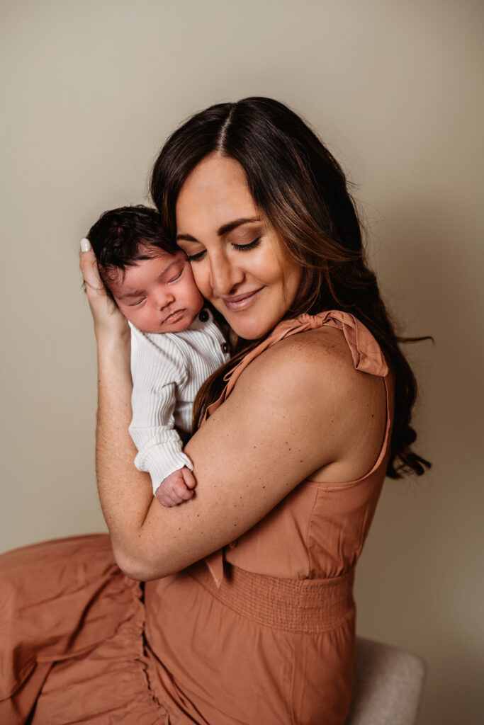 mother in a rusty pink dress sits on a stoll and holds her babies face to her