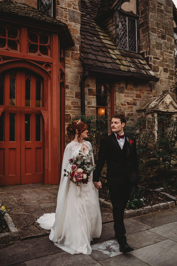 Baltimore wedding photographers photographs game of thrones red wedding theme with bride and groom holding hands and walking through a courtyard at a castle wedding venue with bride in a cape and holding a red rose bouquet