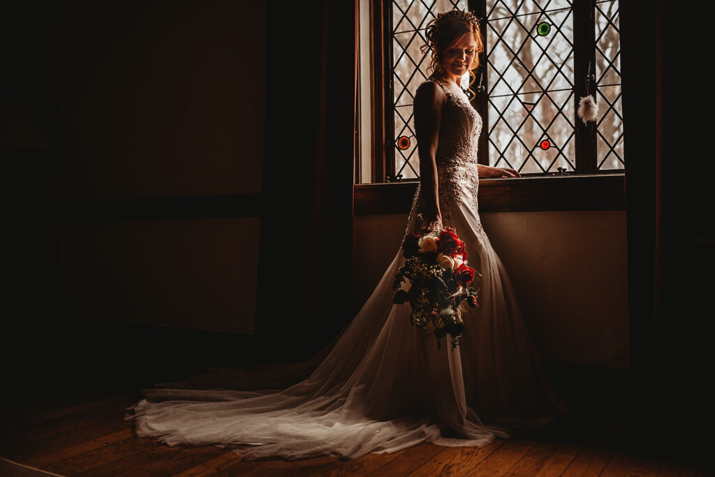 bridal portraits in a bridal suite at castle wedding venue in Maryland with bride holdin gher red rose bouquet to her side as she looks out old stain glass window