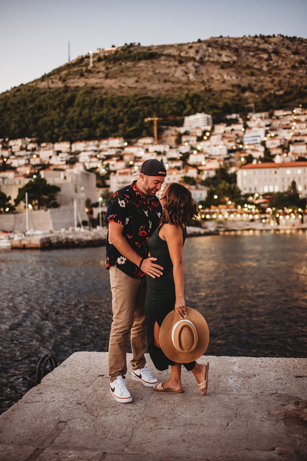Destination Engagement Photos in Croatia with man and woman embracing on a pier at golden hour with a mountain landscape covered in small white cottages