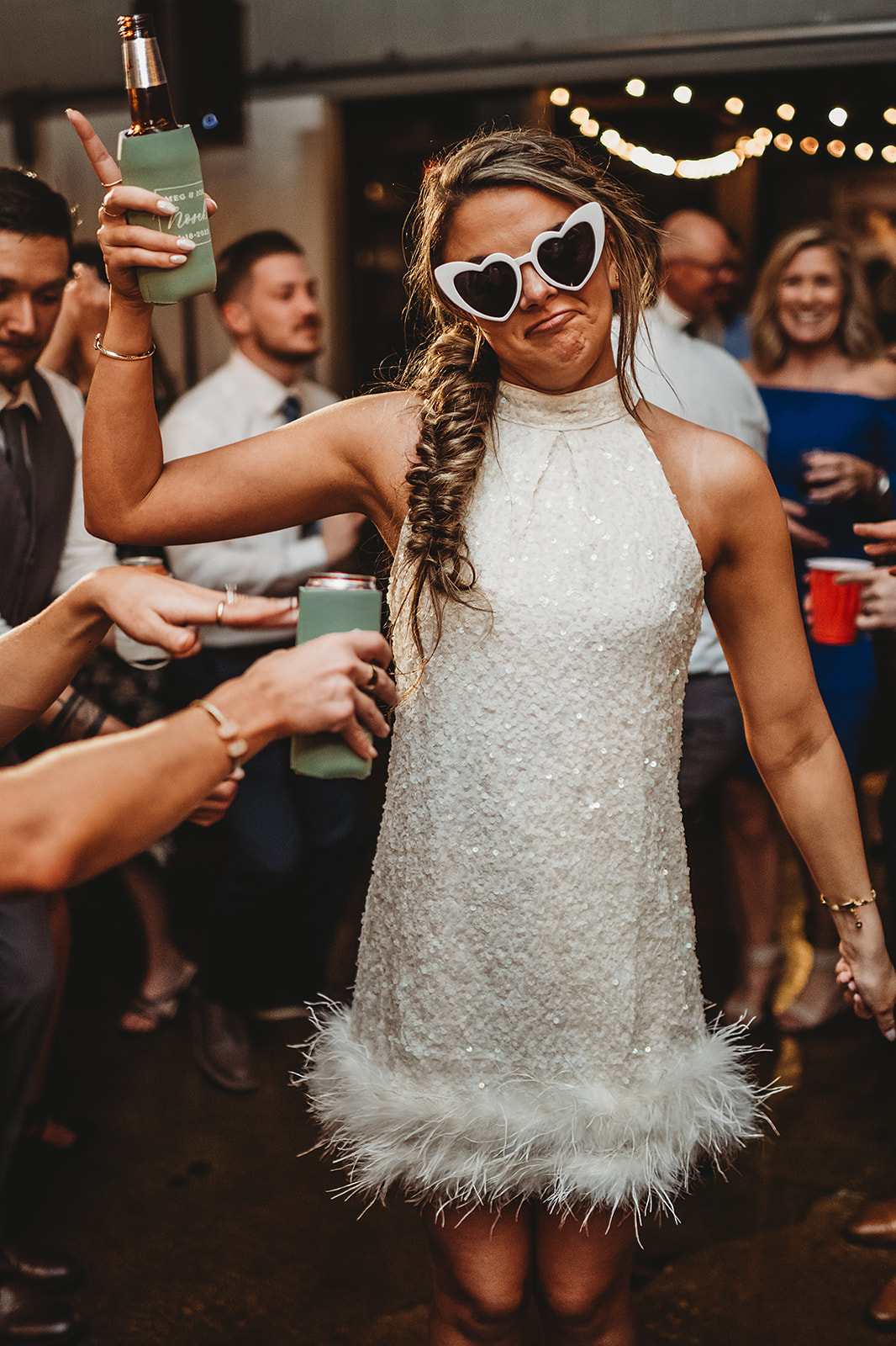 bride in her wedding reception dress holding a drink in one hand and smiling while wearing white heart shaped glasses captured by Baltimore photographers