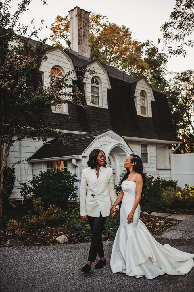 Baltimore photographers captures two brides holding hands and walking outside of their Baltimore wedding venue as they smile at one another 