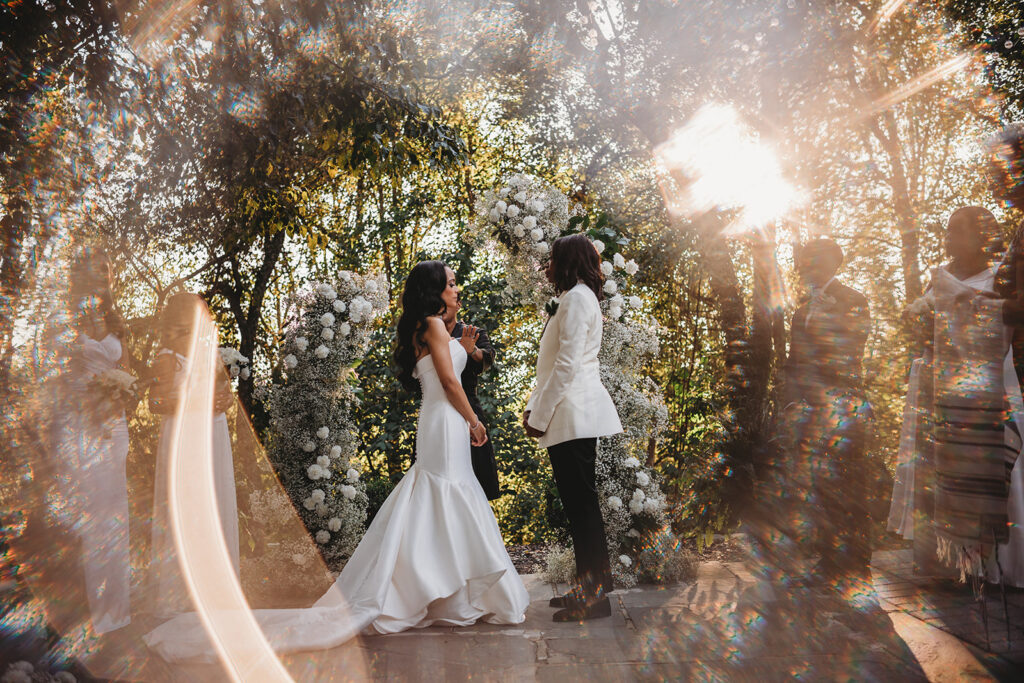 lgbtq wedding with lesbian couple standing together during their ceremony in the woods with the light shining through the leaves of the trees behind them 