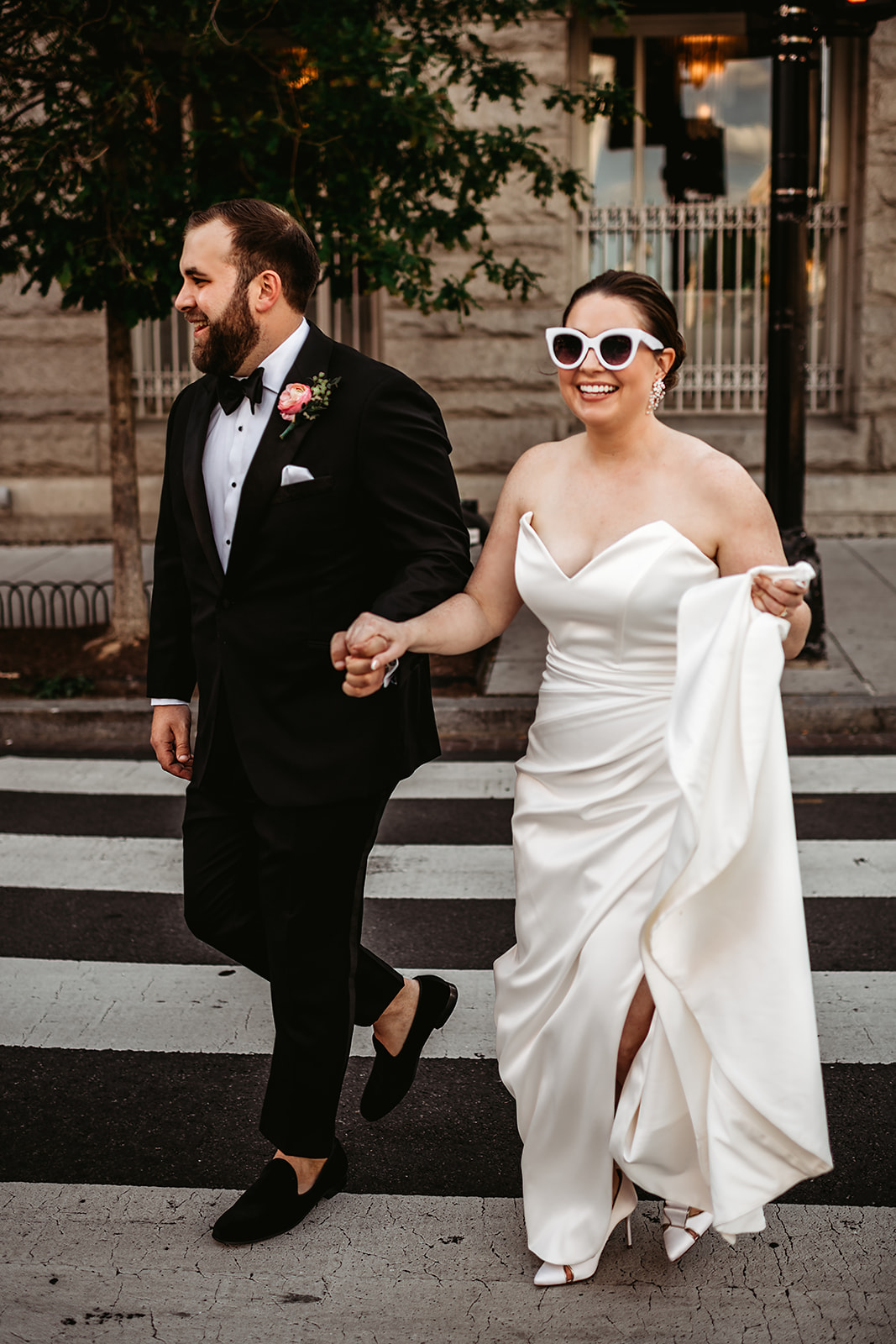 bride and groom holding hands as they cross a street together with the bride in white heart sunglasses holding her dress from dragging on the floor and smiling really big