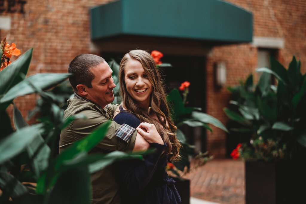 Baltimore wedding photographers capture man hugging woman in front of big plants during Fells Point engagement photos