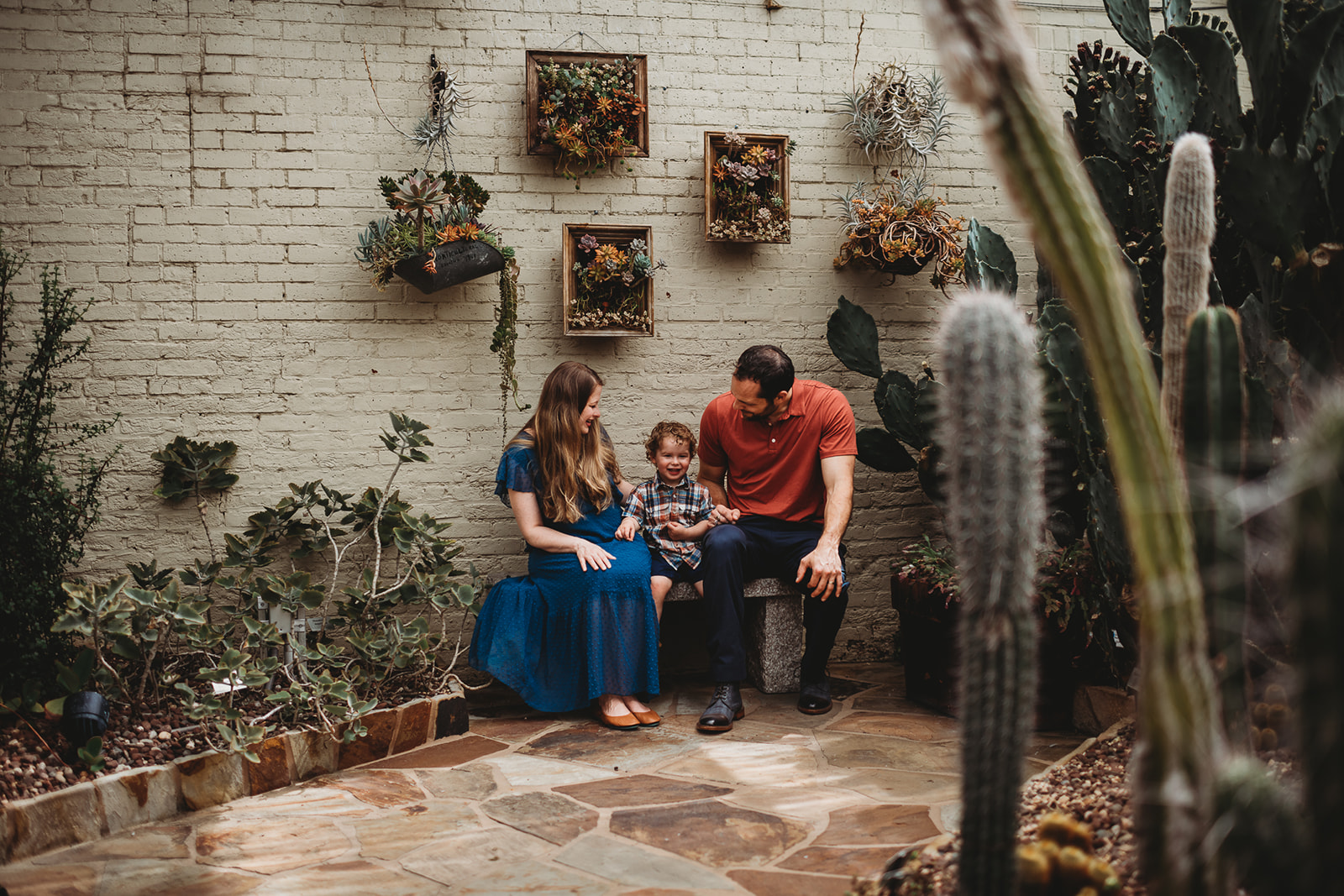 mother and father sitting on a bench together with their toddler child in a courtyard full of cacti