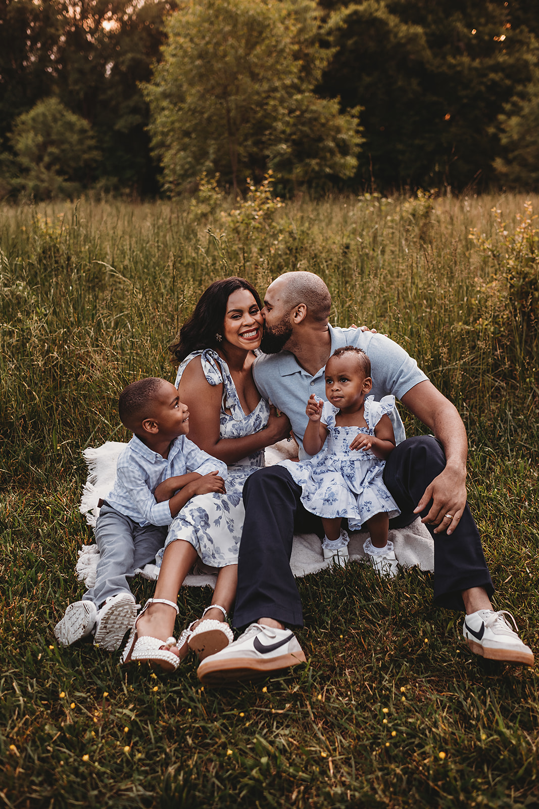 Maryland family photographer captures outdoor family picture with young family sitting together on a blanket at sunset with the father kissing the mother on the cheek while the children sitting on their laps