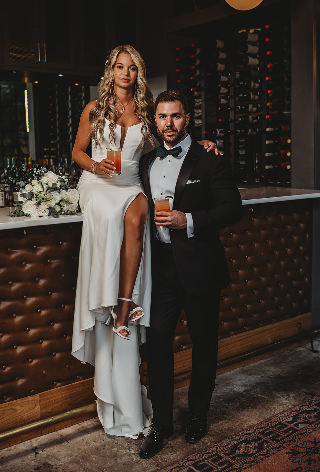 baltimore wedding photographers captures bride sitting on a bar at the wool factory holding a drink while her groom stands beside her and holds her waist