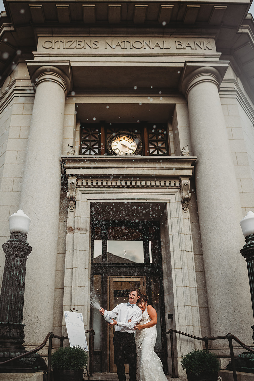 groom pops a bottle of champagne in front of Citizens ballroom on the stairs as the bride laughs and ducks behind the groom