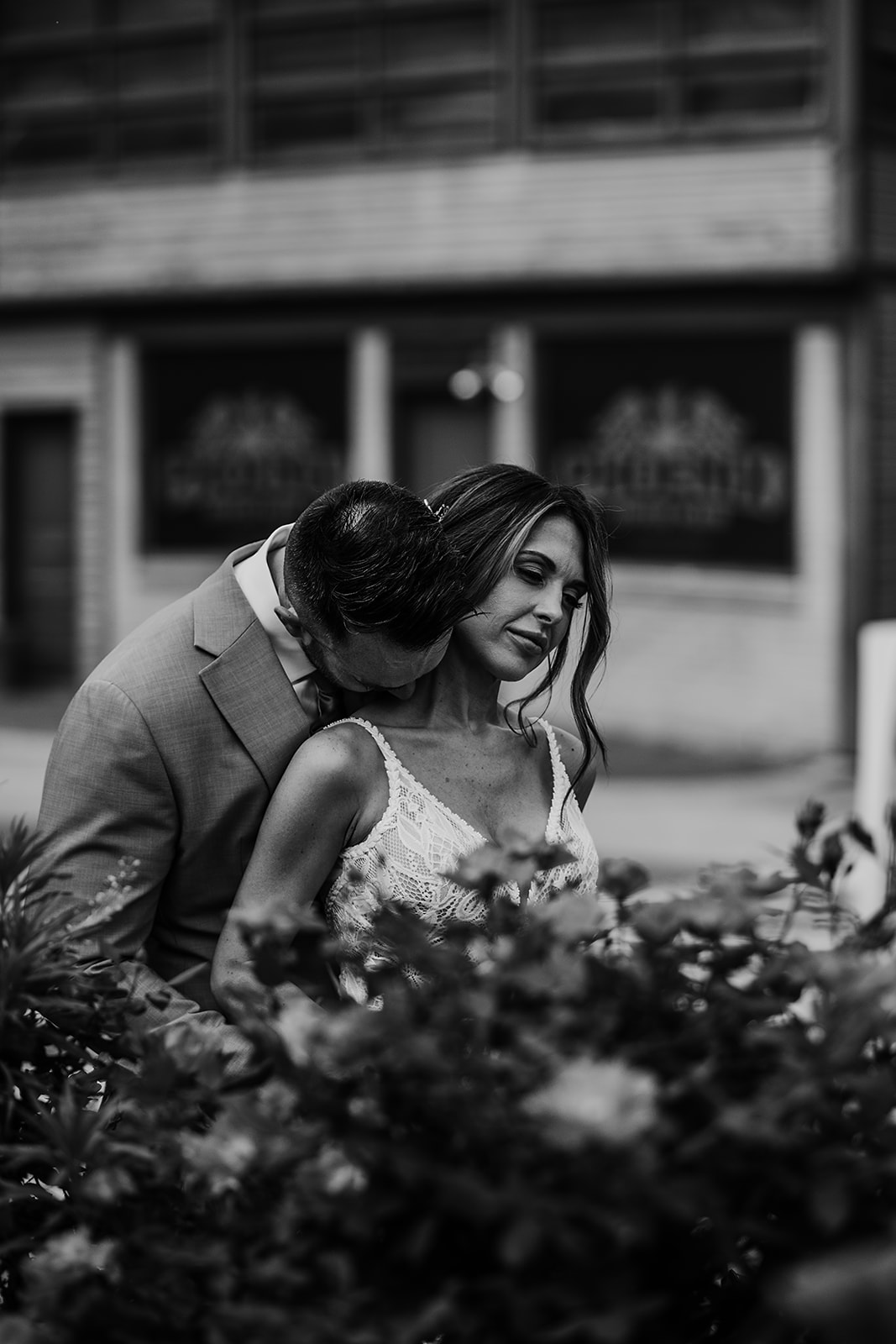 A timeless black-and-white portrait of a bride and groom sharing a quiet, intimate moment. The groom leans in to kiss the bride’s shoulder as she closes her eyes, embracing the connection between them. The delicate details of her lace gown and the soft blur of nearby flowers add an elegant touch, while the urban backdrop grounds the scene in authenticity. Captured by a Baltimore wedding photographer, this image is a beautiful reflection of love and tenderness.