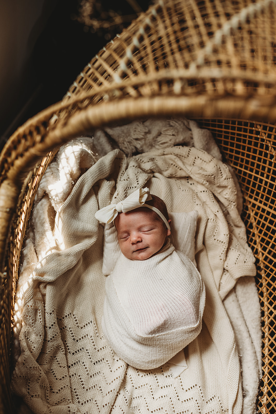 A serene newborn portrait captured in a Baltimore newborn photographer studio, featuring a baby swaddled in a soft cream wrap and nestled in a wicker bassinet lined with textured, knitted blankets. The baby wears a simple cream bow headband and appears to be smiling gently while sleeping. The warm, natural light filtering through the bassinet enhances the cozy, timeless feel of the scene.