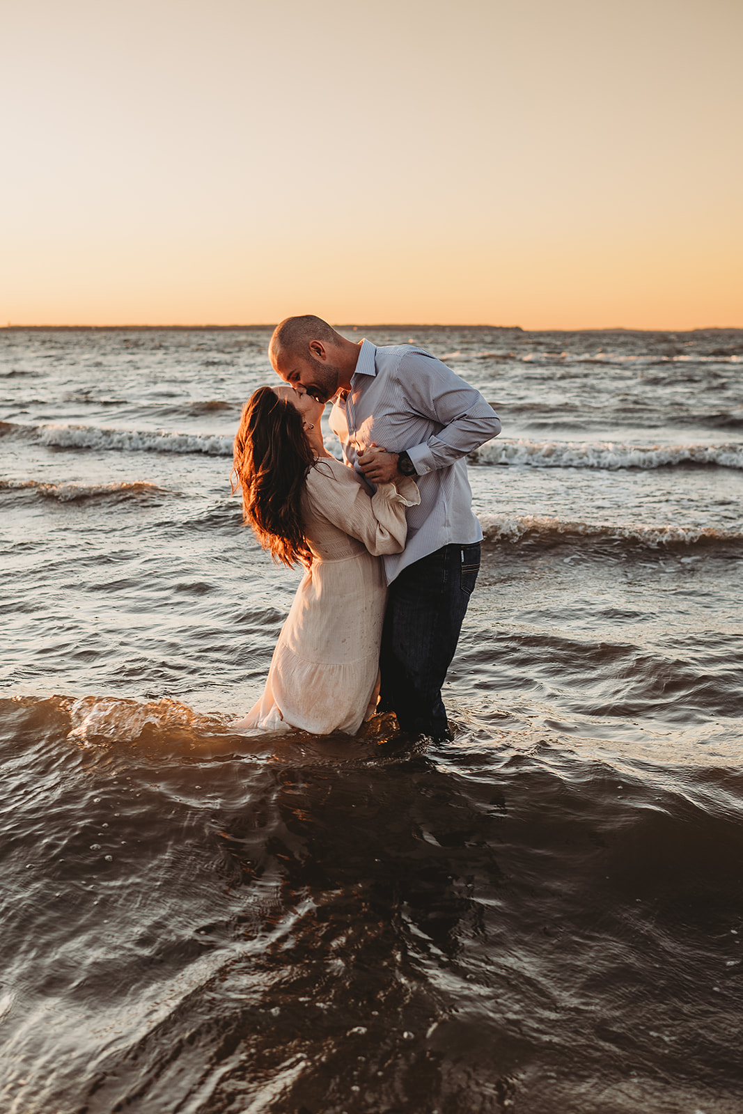 A couple standing in the waves at sunset, holding each other closely as the man leans in to kiss the woman’s forehead, featured in a Baltimore engagement photos session by a Baltimore photographer.