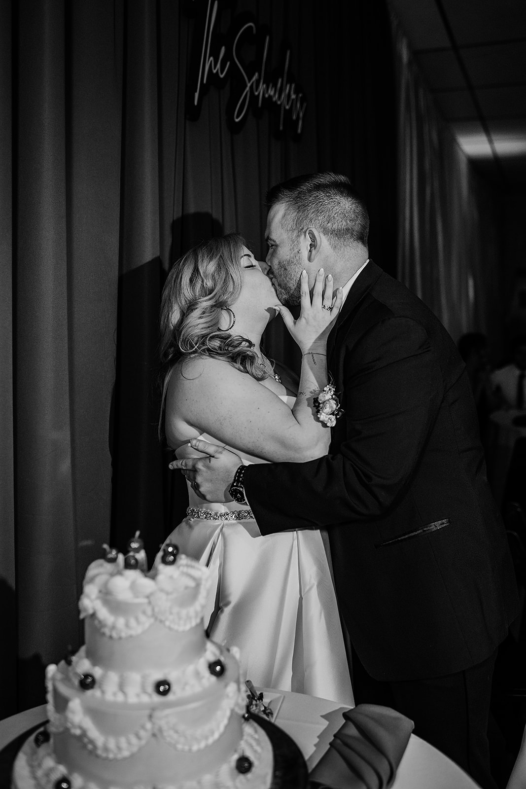 Black and white photo of bride and groom kissing in front of their wedding cake, with "The Schuelers" sign in the background, captured during a Locust Point wedding by Baltimore wedding photographer.