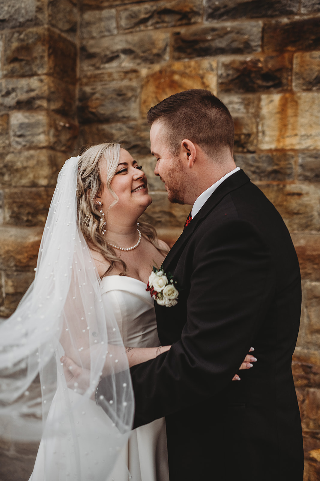 Bride and groom embracing and smiling at each other in front of a rustic stone wall on their wedding day, with the bride in a strapless white dress and veil and the groom in a black suit with a white and red boutonniere.