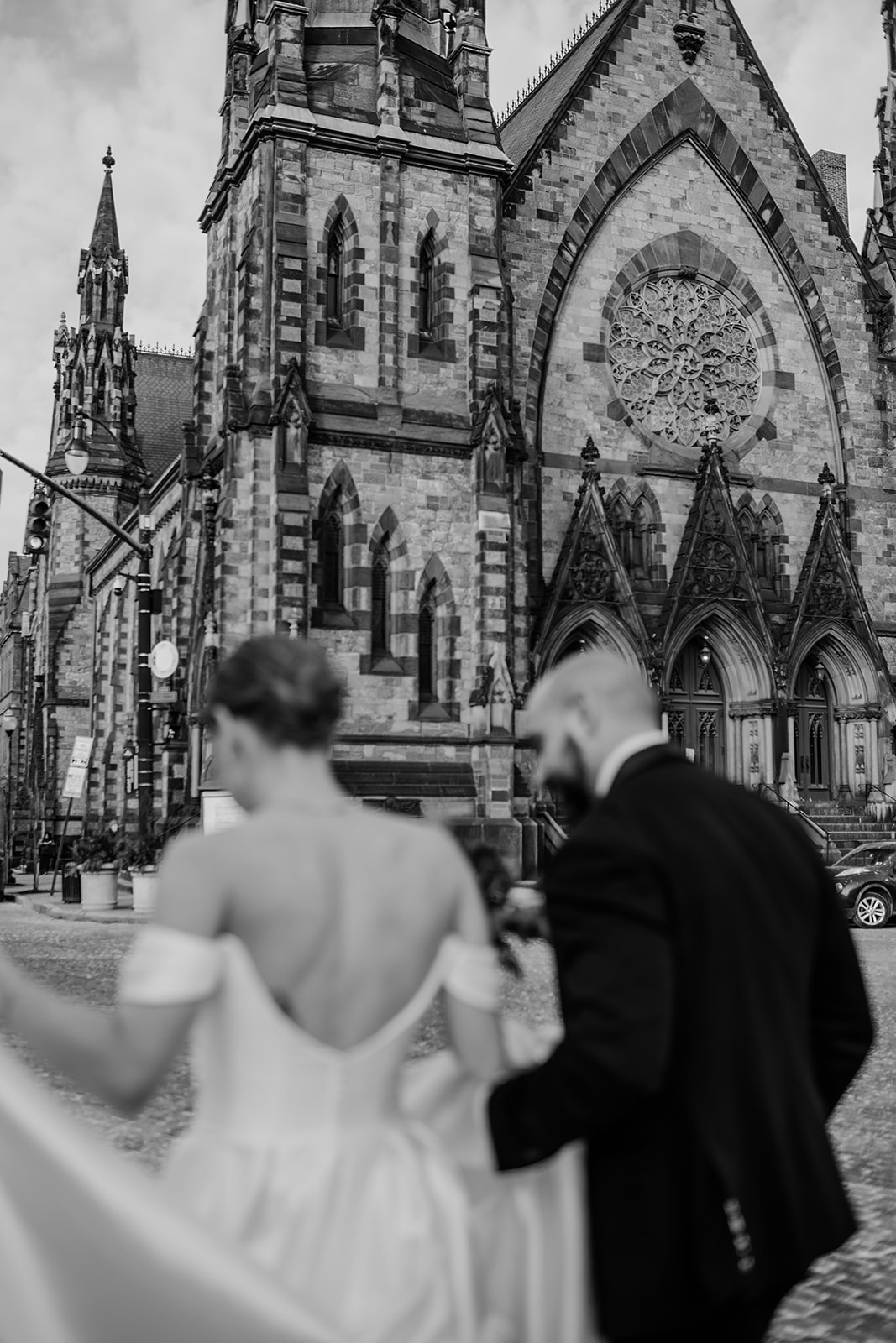 Black and white photo of a bride and groom walking hand in hand toward the historic Gothic-style Mount Vernon Place United Methodist Church in Baltimore, its detailed stone facade and rose window in the background.