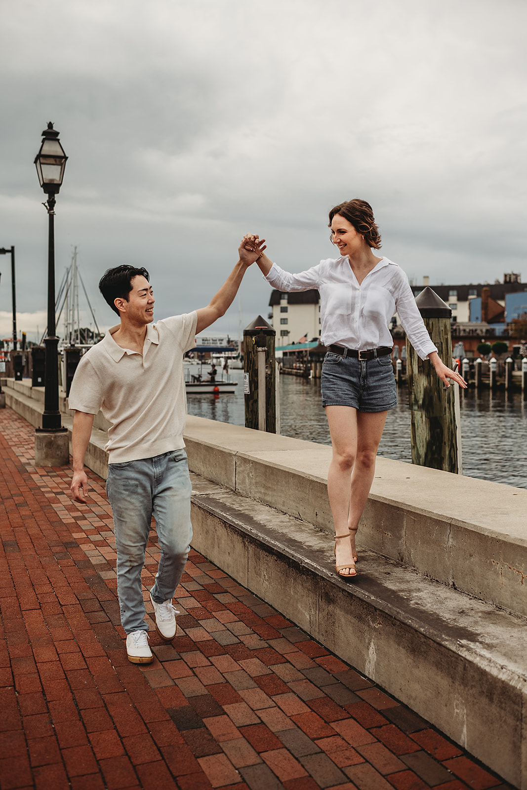 Engaged couple walking along the Annapolis waterfront, with the woman balancing on a ledge while holding hands with her partner, photographed by a Baltimore photographer during their Downtown Annapolis, MD engagement session.