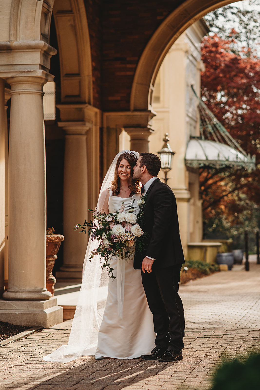 Bride holding her bouquet and smiling as the groom kisses her temple under the stone archways of Evergreen Museum