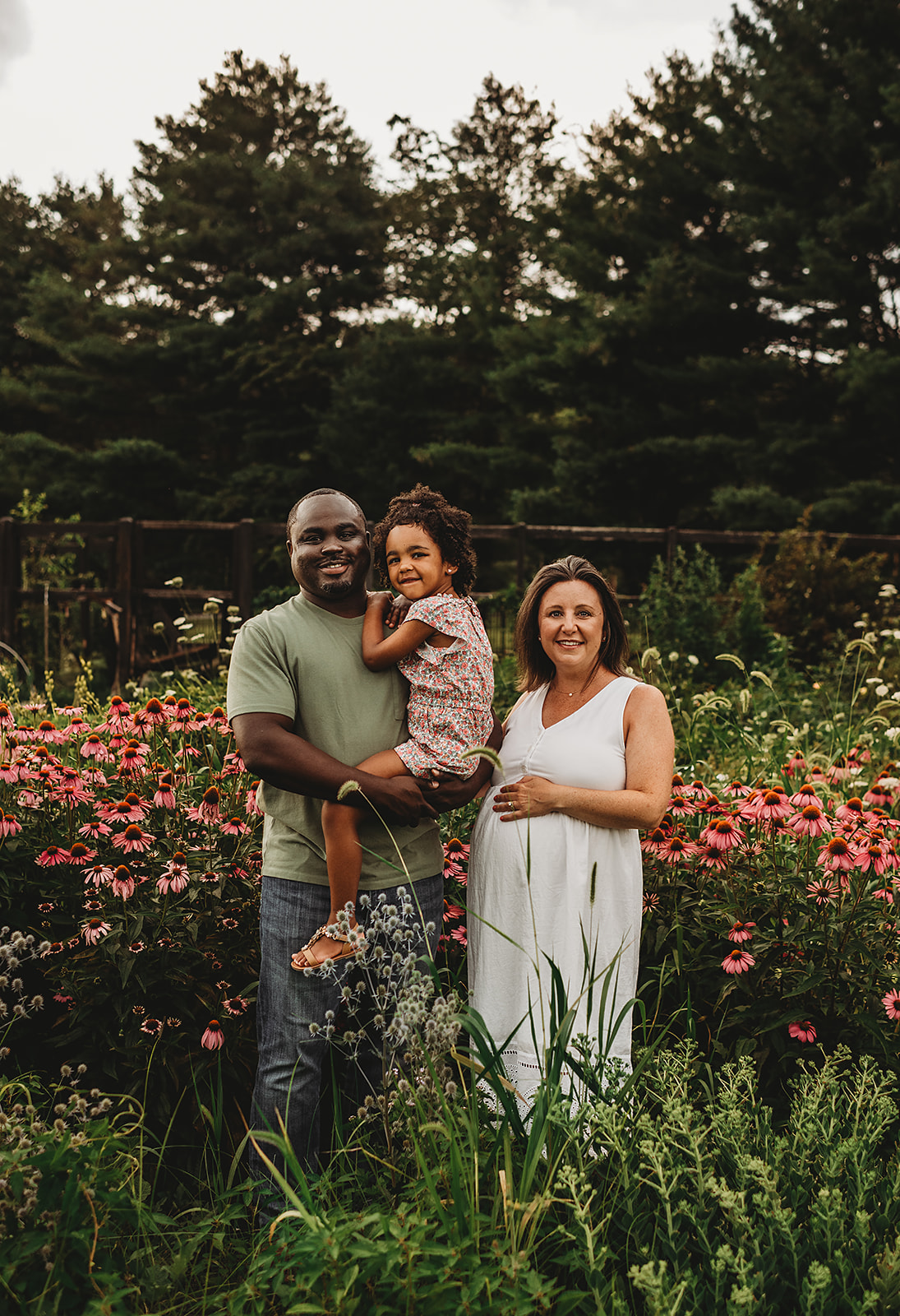 Family of three stands together in a wildflower field filled with pink coneflowers, with the father holding their daughter and the mother cradling her baby bump, captured during Wildflower Photos in Maryland by a Baltimore photographer