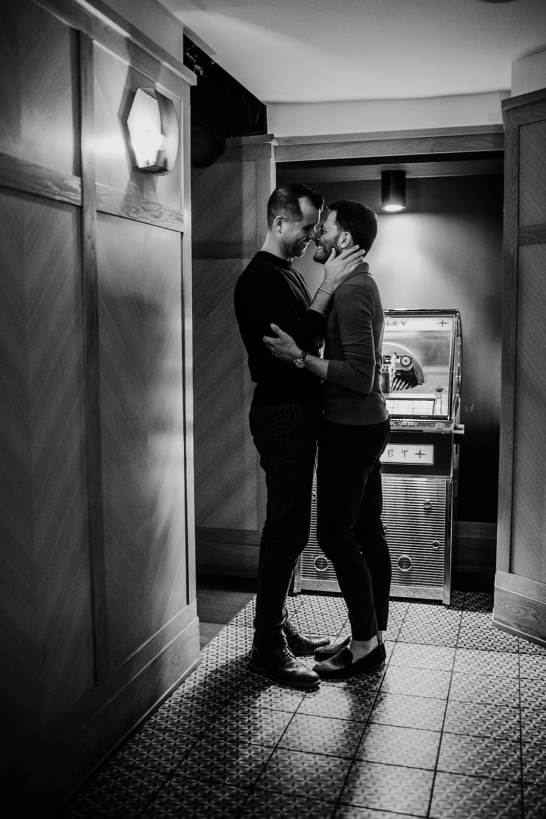 Two men stand closely in a hallway beside a vintage jukebox at Revival Hotel, holding each other and smiling during their hotel engagement photos by a Baltimore wedding photographer.