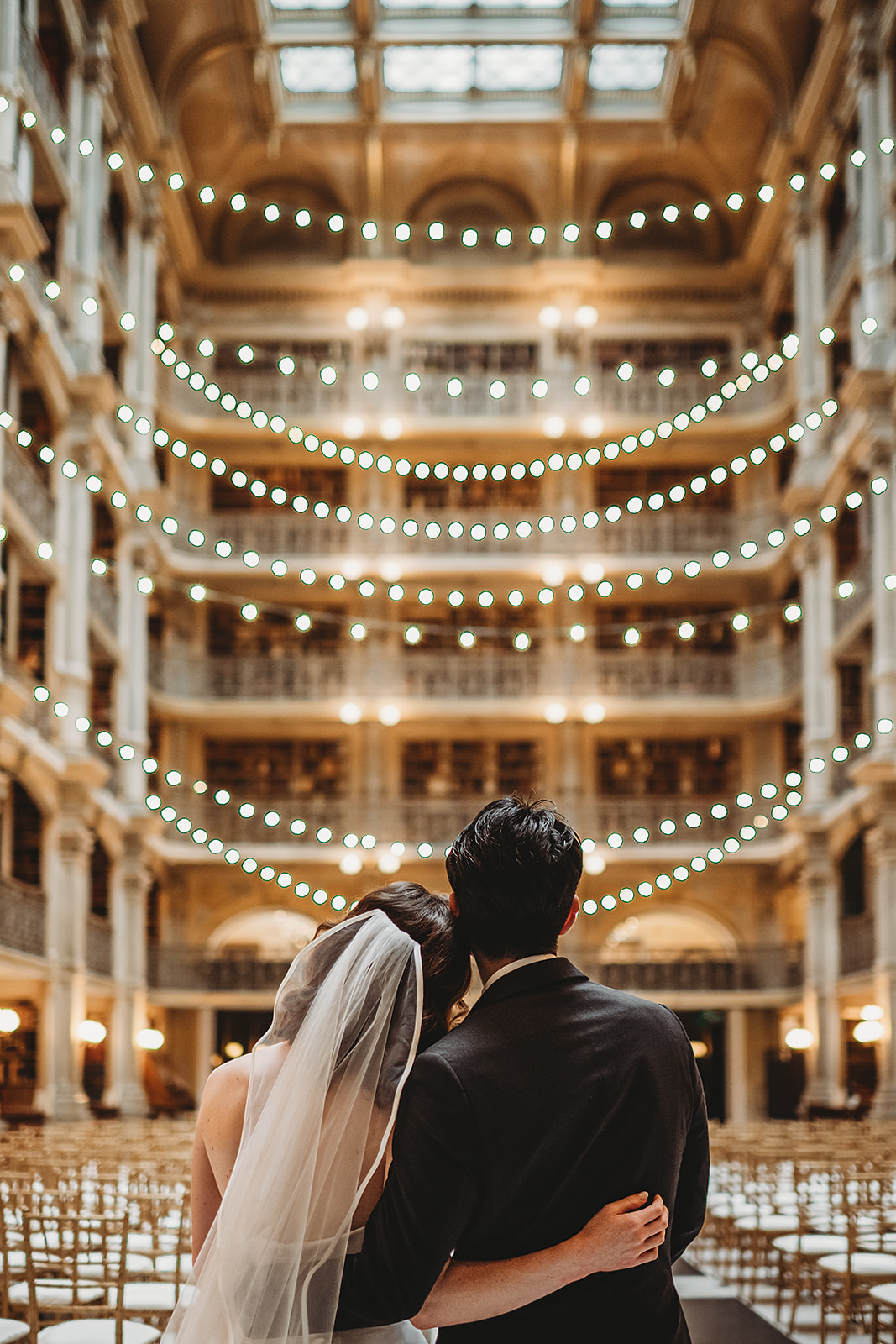 Newlywed couple stands arm in arm in the George Peabody Library Maryland, gazing up at the multi-level architecture and glowing string lights above, photographed by a Baltimore wedding photographer