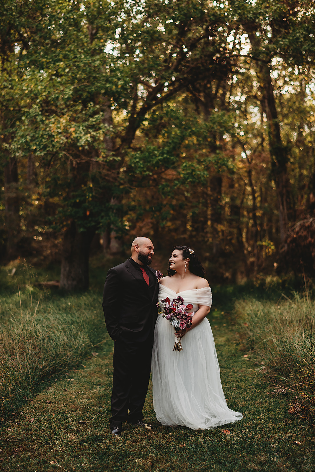 Bride and groom smiling at each other while standing on a wooded path, with the bride holding a jewel-toned bouquet, taken during a fall wedding at Patapsco Valley State Park by a Baltimore wedding photographer
